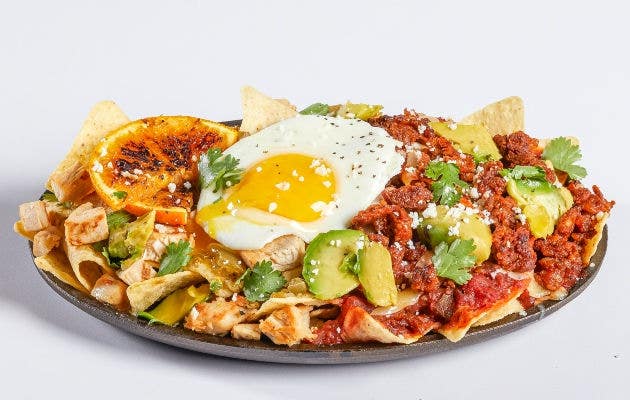 Chilaquiles rojos with chorizo and chilaquiles verdes with pulled chicken, served with avocado and grilled oranges on a dark plate, set against a white background