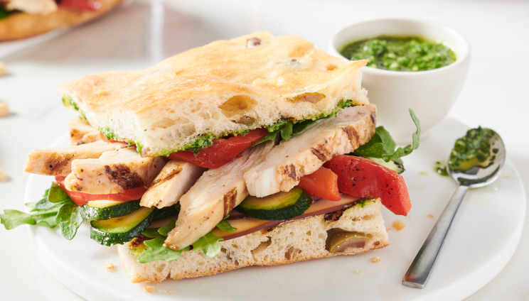 Grilled chicken sandwich with fresh vegetables and pesto sauce on focaccia bread, served on a plate